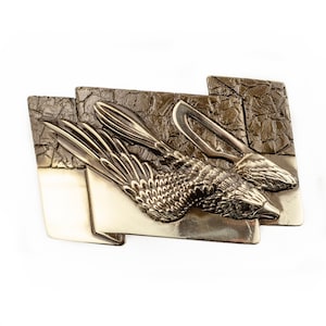 May include: A gold-toned metal brooch with a detailed design of two birds in flight. The birds are depicted with their wings spread and their heads turned towards each other. The brooch has a textured background.