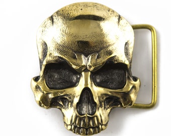 Puede incluir: Una hebilla de cinturón de calavera dorada con un diseño detallado. La hebilla es de metal y tiene forma rectangular.