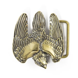 May include: A gold-toned metal belt buckle featuring a detailed eagle design with spread wings. The eagle's head is facing down, and the buckle has a rectangular shape with a smooth, polished finish.