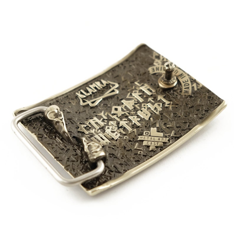 Peut inclure: Boucle de ceinture rectangulaire en m&eacute;tal avec une surface textur&eacute;e gris fonc&eacute;. La boucle porte le mot "KLAMKA" et d'autres textes, avec un cadre argent&eacute; et un tenon m&eacute;tallique pour l'attache. Les mots "METAL ART CAST" sont &eacute;galement visibles.