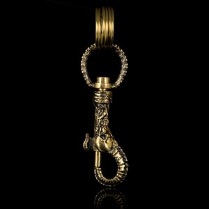 K&ouml;nnte beinhalten: Ein goldfarbener Schl&uuml;sselanh&auml;nger aus Metall mit einem detaillierten, verzierten Design. Der Schl&uuml;sselanh&auml;nger verf&uuml;gt &uuml;ber einen Haken und einen Ring.