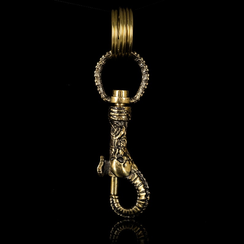 K&ouml;nnte beinhalten: Ein goldfarbener Schl&uuml;sselanh&auml;nger aus Metall mit einem detaillierten, verzierten Design. Der Schl&uuml;sselanh&auml;nger verf&uuml;gt &uuml;ber einen Haken und einen Ring.