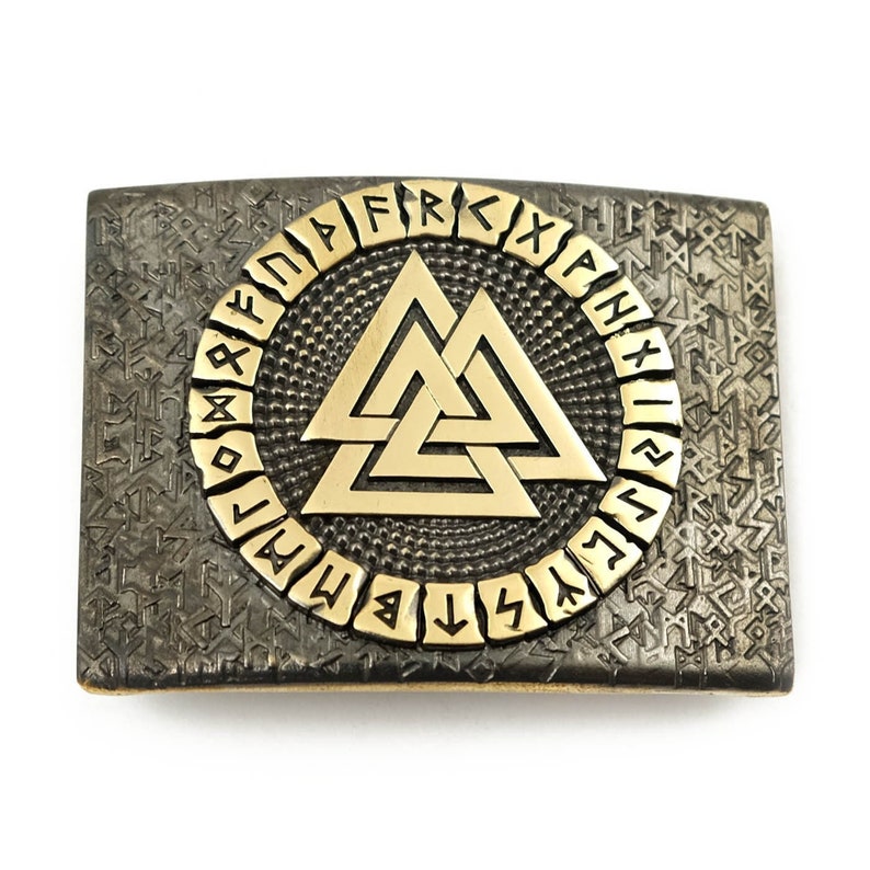 Peut inclure: Boucle de ceinture rectangulaire en m&eacute;tal. Elle pr&eacute;sente un symbole Valknut dor&eacute; au centre, entour&eacute; de runes dor&eacute;es. Le fond est en m&eacute;tal gris fonc&eacute; textur&eacute; avec d'autres symboles runiques.