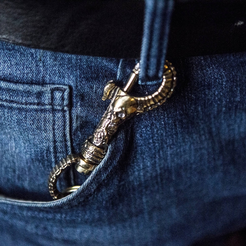 K&ouml;nnte beinhalten: Ein goldfarbener Metallhaken mit einem verzierten Design, der an einer G&uuml;rtelschlaufe einer blauen Jeans befestigt ist.
