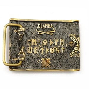 Peut inclure: Boucle de ceinture rectangulaire en m&eacute;tal avec un cadre et des accents dor&eacute;s. La boucle pr&eacute;sente un fond gris fonc&eacute; avec des lettres et des symboles dor&eacute;s. Le texte "KLAMKA" est en haut et "MADE IN UKRAINE" sur le c&ocirc;t&eacute; droit.