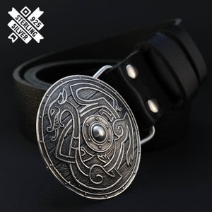 Puede incluir: Una hebilla de cintur&oacute;n de plata con un dise&ntilde;o detallado de un escudo con un drag&oacute;n. La hebilla est&aacute; unida a un cintur&oacute;n de cuero negro.