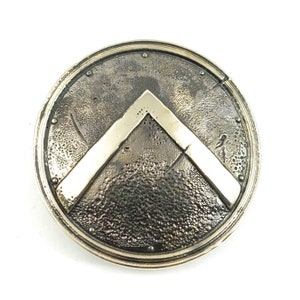 Peut inclure: Un bouclier rond argenté avec un motif en chevron blanc stylisé en relief. Le bouclier a une surface texturée.