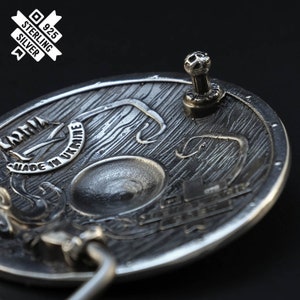 Puede incluir: Una hebilla de cintur&oacute;n de plata con un dise&ntilde;o detallado de un escudo con textura de madera. La hebilla est&aacute; marcada con "925 Sterling Silver" y "Made in Ukraine".