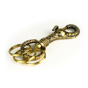 K&ouml;nnte beinhalten: Ein goldfarbener Schl&uuml;sselanh&auml;nger mit einem detaillierten Sch&auml;del-Design und mehreren ineinandergreifenden Ringen.