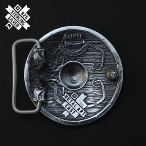 Puede incluir: Una hebilla de cintur&oacute;n de plata con un dise&ntilde;o detallado de un escudo con un kraken y el texto "KASHMA MADE IN UKRAINE METAL ART CAST 925 STERLING SILVER".