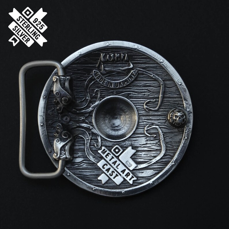 Puede incluir: Una hebilla de cintur&oacute;n de plata con un dise&ntilde;o detallado de un escudo con un kraken y el texto "KASHMA MADE IN UKRAINE METAL ART CAST 925 STERLING SILVER".