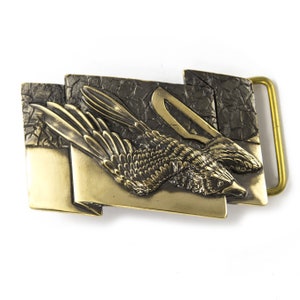 May include: A gold-toned belt buckle with a detailed design of two birds in flight. The buckle has a textured, almost leather-like surface.