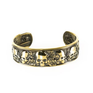Pode incluir: Uma pulseira de metal dourado com um design detalhado de caveiras e rosas. A pulseira é aberta e tem uma borda lisa e arredondada.