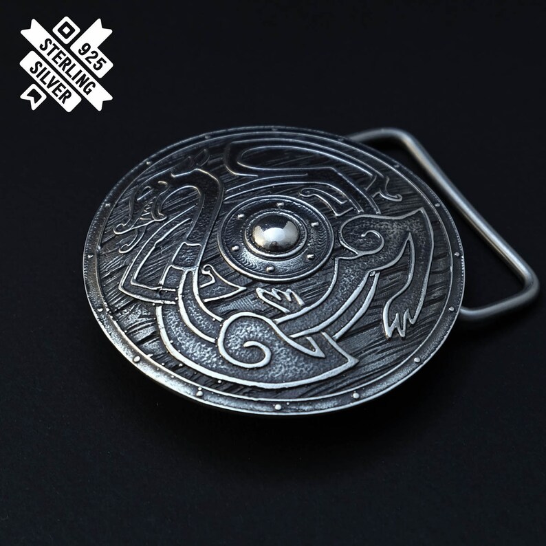 Puede incluir: Una hebilla de cintur&oacute;n de plata con un dise&ntilde;o circular que presenta un escudo con un motivo de drag&oacute;n. La hebilla est&aacute; hecha de plata de ley y tiene un sello de 925.