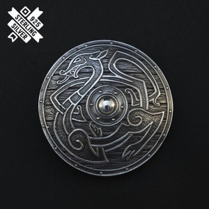 Puede incluir: Un colgante de escudo de plata con un dise&ntilde;o detallado de un drag&oacute;n. El escudo es redondo y tiene un jefe central elevado. El colgante est&aacute; marcado con "925 Sterling Silver".