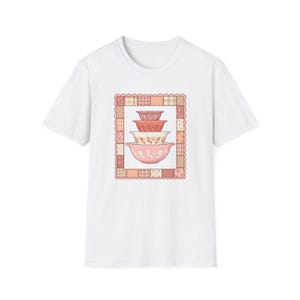May include: White t-shirt featuring a graphic of four stacked mixing bowls in shades of pink and red, with floral patterns. The bowls are set within a decorative, patchwork-style frame.