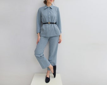 May include: A blue and white gingham patterned pantsuit with a collared shirt and long sleeves. The pantsuit has a black belt with a gold buckle.