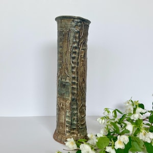May include: A tall, textured ceramic vase with a cylindrical shape. The vase features a mottled brown and green color scheme with intricate carved details. The vase is approximately 12 inches tall and has a unique, artistic design, suitable for home decor.