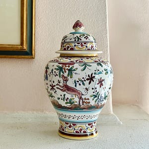 Peut inclure: Un vase en céramique blanc avec couvercle, orné d'un motif peint à la main représentant un chien marron, des fleurs et du feuillage. Le vase a une forme arrondie avec un motif coloré et détaillé et un petit bouton sur le couvercle.