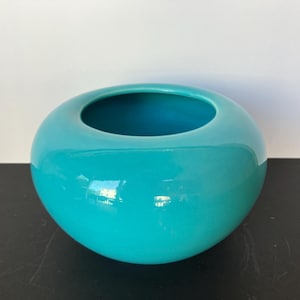 Puede incluir: Un cuenco de cerámica turquesa con una amplia abertura. El cuenco tiene un acabado liso y brillante y una forma redondeada. El interior del cuenco es de un tono turquesa más oscuro. El cuenco está sobre una superficie oscura.