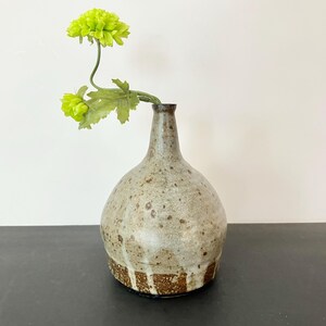 Könnte beinhalten: Eine Keramikvase mit bauchigem Körper und engem Hals, die eine gesprenkelte beige-graue Glasur aufweist, welche in einen dunkelbraunen, texturierten Boden übergeht. Ein einzelner Stiel mit zwei leuchtend lindgrünen Chrysanthemenblüten steckt darin.