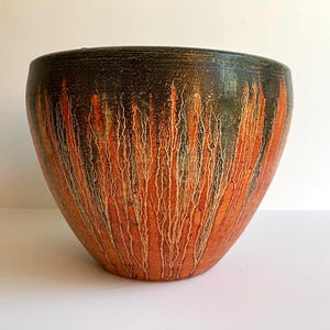 May include: A rounded ceramic bowl featuring a dark charcoal black rim that transitions into a vibrant orange base. The surface displays a textured, vertical drip glaze pattern in cream and white, creating a stark contrast against the dark colors.