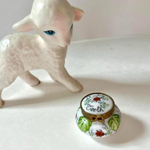May include: A white ceramic lamb figurine with blue eyes positioned next to a small, round, white porcelain trinket box with a brass hinge. The lid features a hand-painted ladybird and the text Baby Teeth, with green leaves and ladybirds below.
