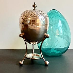 May include: A silver-toned metal egg-shaped container with a decorative leaf-patterned lid and a small finial, supported by a three-legged stand with ball feet. Beside it sits a large, translucent, teardrop-shaped teal glass decorative object.