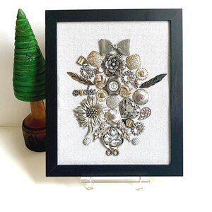 Könnte beinhalten: Ein gerahmtes Wandkunstwerk mit einer Collage aus verschiedenen silberfarbenen Vintage-Schmuckstücken, Knöpfen und einem kleinen Zifferblatt auf hellgrauem Stoffhintergrund. Der schwarze rechteckige Rahmen umschließt die Sammlung metallischer Objekte.
