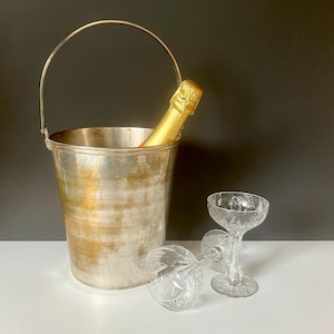 May include: A silver champagne bucket with a handle holds a gold-wrapped bottle. Two clear, etched champagne glasses are next to the bucket. One glass is upright, and the other is lying on its side. The background is dark.