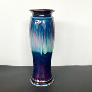 May include: A tall, glazed ceramic vase with a flared base and a slightly wider top. The vase features a blend of colors, including deep blues, purples, and hints of green and pink, creating a flowing, artistic effect. The rim and base have a dark brown accent.
