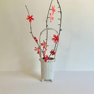 Op de afbeelding: Een witte keramische vaas met vlechtwerkpatroon, voorzien van een hoog, gebogen handvat en drie kleine voetjes. De vaas bevat diverse dunne, donkerbruine takken versierd met felroze, vijfbladige kunstbloemen die omhoog steken.