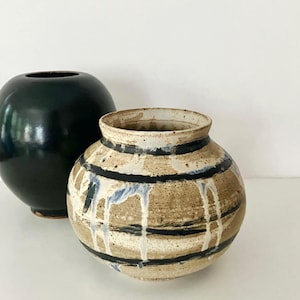Könnte beinhalten: Zwei Keramikvasen. Eine Vase ist einfarbig, glänzend dunkelgrün und hat eine abgerundete, kugelförmige Gestalt. Die zweite Vase besteht aus texturierter, hellbeiger Keramik mit horizontalen schwarzen Streifen und weißen Glasurtropfen.
