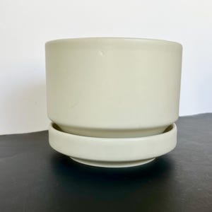 Puede incluir: Una maceta de cerámica beige claro con un platillo a juego. La maceta cilíndrica se asienta sobre una base baja y redonda. La maceta es adecuada para uso en interiores o exteriores. El color neutro complementa varios estilos de decoración.