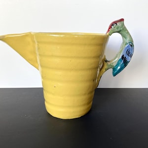 May include: A yellow ceramic pitcher with a bird-shaped handle. The pitcher has a spout and a ribbed texture. The handle is painted with red, green, and blue details. The pitcher is likely used for pouring liquids.