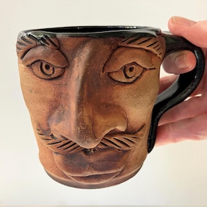 May include: A brown ceramic mug with a sculpted face design. The mug features a detailed face with eyes, a nose, and a mustache. The handle is black, and the interior is also black.