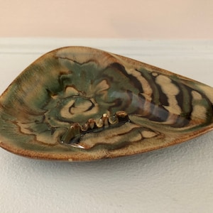 Könnte beinhalten: Ein dreieckiger Aschenbecher aus Keramik mit brauner, grüner und weißer Glasur. Der Aschenbecher hat ein wellenförmiges, abstraktes Design.