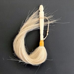 May include: A cream-colored carved figure handle attached to a long, flowing bundle of light-colored horsehair. The handle features a stylized human form with a small loop cord and a distinct yellow band connecting the carving to the hair fibers.
