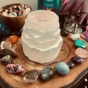 Può includere: Un portacandele in selenite bianca con un'apertura circolare nella parte superiore, circondato da una varietà di cristalli colorati su una fetta di legno. I cristalli includono ametista, quarzo rosa, quarzo trasparente e malachite.