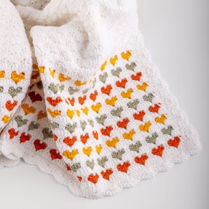 crochet baby heart blanket pattern with white yarn as base color in the middle with the shell stitch and autumn colors green orange and red for the hearts