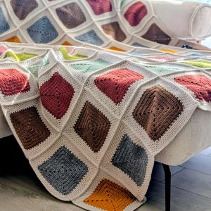 May include: A handmade crochet blanket draped over a sofa. The blanket features a repeating pattern of colorful squares in shades of red, brown, blue, and yellow, set against a cream-colored background. The blanket is a cozy home decor item.