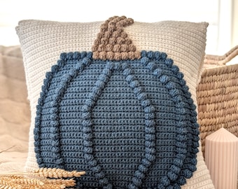 May include: A cream-coloured square cushion featuring a large, textured, blue crocheted pumpkin design. The pumpkin has a brown stem. The cushion is on a soft, neutral-coloured surface, with a candle and wheat stalks nearby.