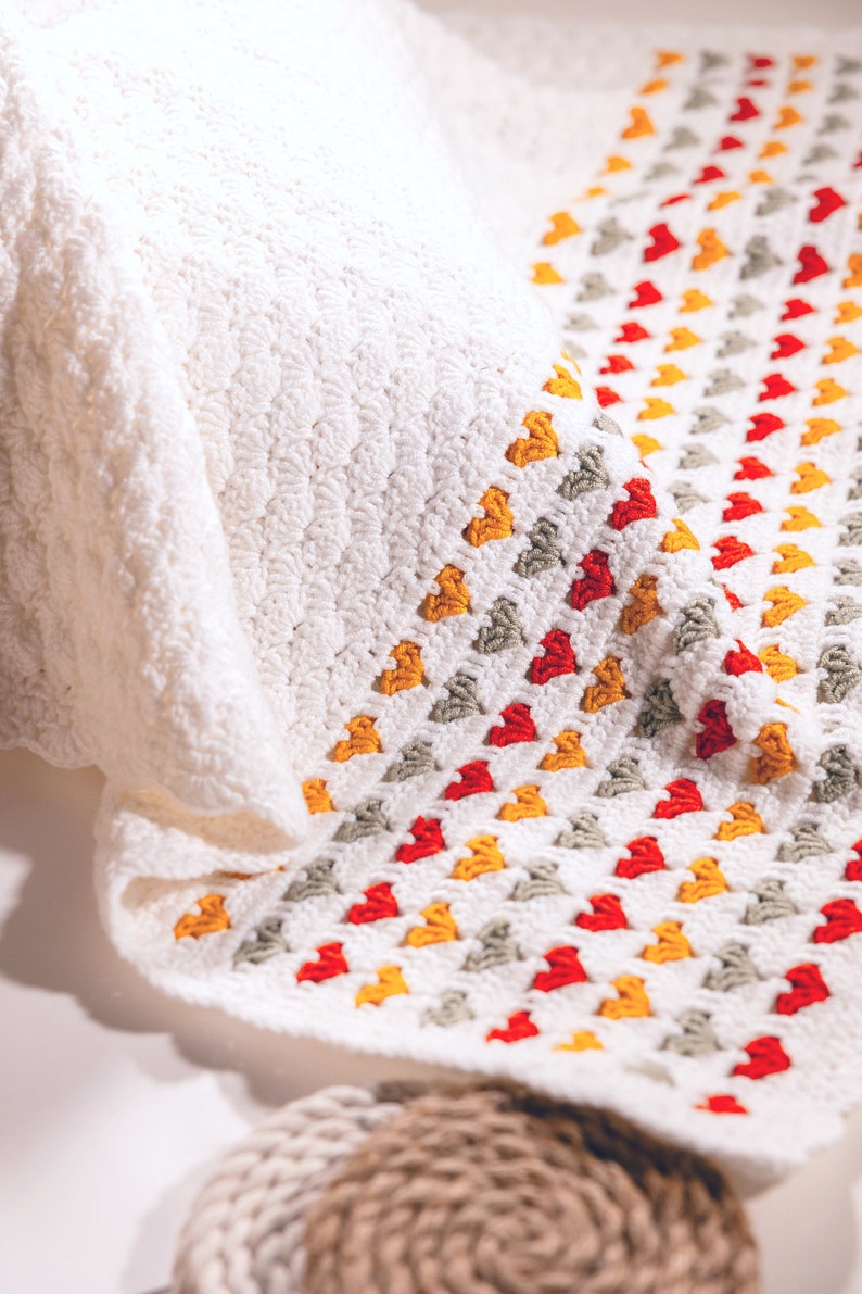 crochet baby heart blanket pattern with white yarn as base color in the middle with the shell stitch and autumn colors green orange and red for the hearts