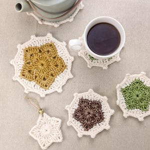 May include: Five crocheted coasters in various colours, including green, brown, and yellow. The coasters are shaped like stars and have a white border. A cup of coffee sits on one of the coasters.