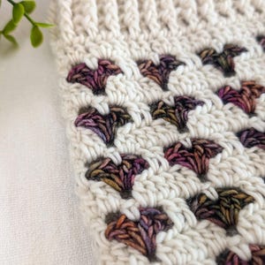 May include: Close-up of a crocheted fabric with a white background and a repeating pattern of small, colorful hearts. The hearts are made with a variegated yarn in shades of brown, purple, and green.