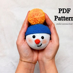 May include: A crocheted snowman head with a blue hat and an orange pom-pom. The snowman has a white face with black eyes, a red nose, and a smiling mouth. The text "PDF Pattern crochet snowman head" is visible in the image.