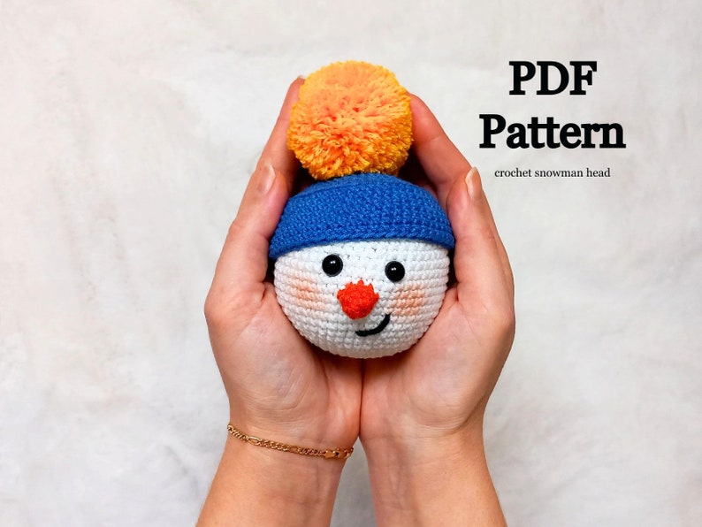 May include: A crocheted snowman head with a blue hat and an orange pom-pom. The snowman has a white face with black eyes, a red nose, and a smiling mouth. The text "PDF Pattern crochet snowman head" is visible in the image.