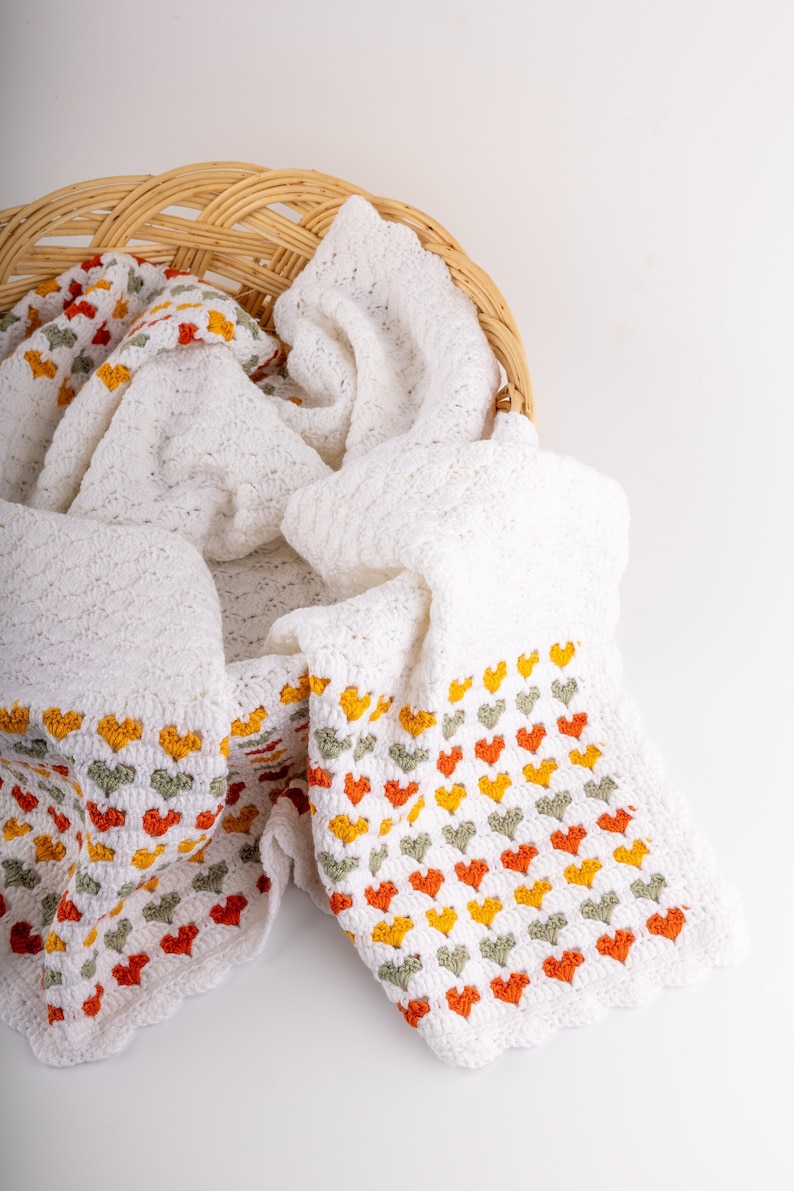 crochet baby heart blanket pattern with white yarn as base color in the middle with the shell stitch and autumn colors green orange and red for the hearts