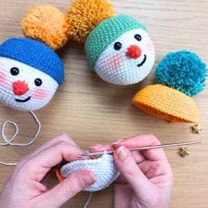 May include: A close-up of a person crocheting a white snowman ornament. The ornament is being worked on with a crochet hook and yarn. There are two other completed snowman ornaments in the background, one with a blue hat and one with a green hat.