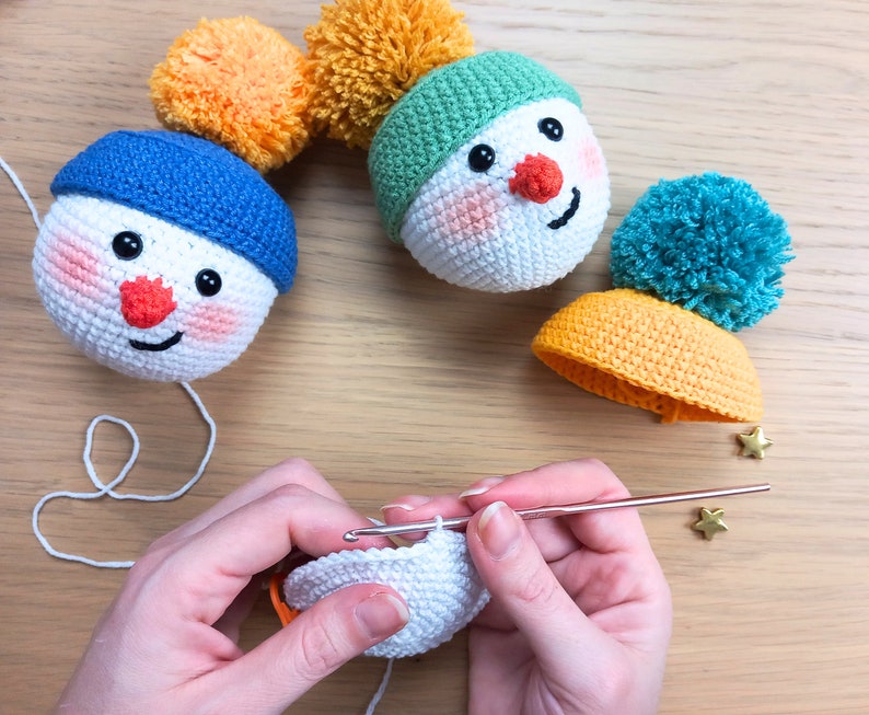 May include: A close-up of a person crocheting a white snowman ornament. The ornament is being worked on with a crochet hook and yarn. There are two other completed snowman ornaments in the background, one with a blue hat and one with a green hat.